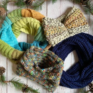 Four knitted scarves in various colors and patterns are arranged on a white wooden background adorned with pine branches and cones.