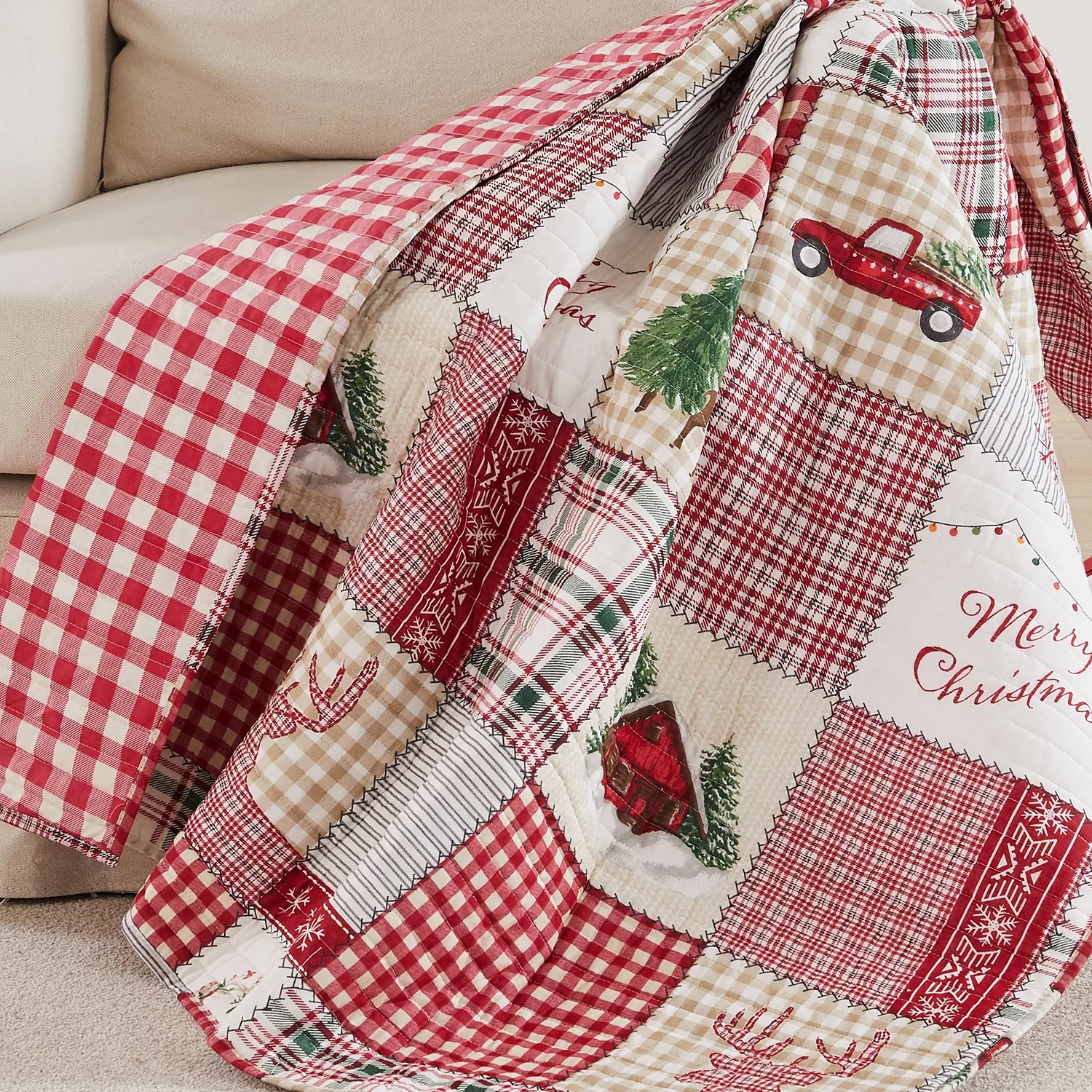 A Christmas-themed quilt features red and beige patchwork with checkered patterns, festive elements like a truck carrying a tree, reindeer, snowflakes, and \“Merry Christmas\“ text.