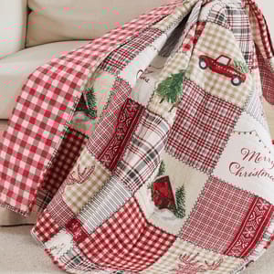 A Christmas-themed quilt features red and beige patchwork with checkered patterns, festive elements like a truck carrying a tree, reindeer, snowflakes, and \“Merry Christmas\“ text.