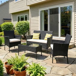 Outdoor patio set with black wicker furniture, including a loveseat, two chairs, and a glass-top coffee table, accented with yellow-striped pillows. Terracotta pots with greenery surround the seating area, set on a stone patio against a house with sliding glass doors.