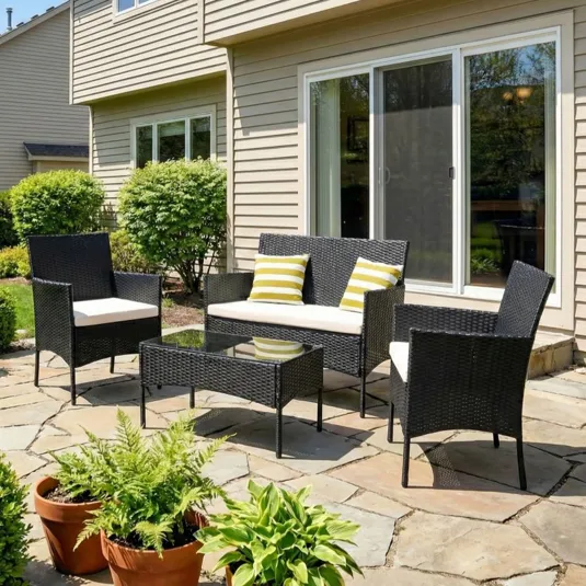 Outdoor patio set with black wicker furniture, including a loveseat, two chairs, and a glass-top coffee table, accented with yellow-striped pillows. Terracotta pots with greenery surround the seating area, set on a stone patio against a house with sliding glass doors.