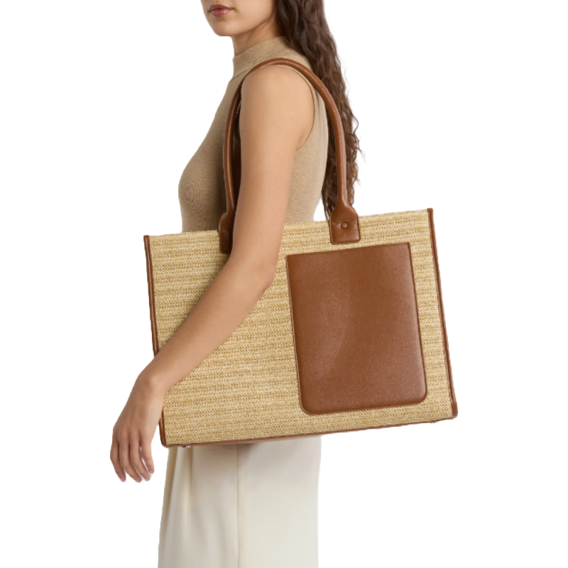 A woman is holding a large rectangular woven tote bag with brown leather handles and a front pocket. She is dressed in a sleeveless beige top and white pants.