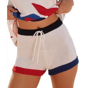 White knit shorts with a black waistband and tie, featuring red and blue trim at the leg openings; paired with a red, white, and blue patterned top.