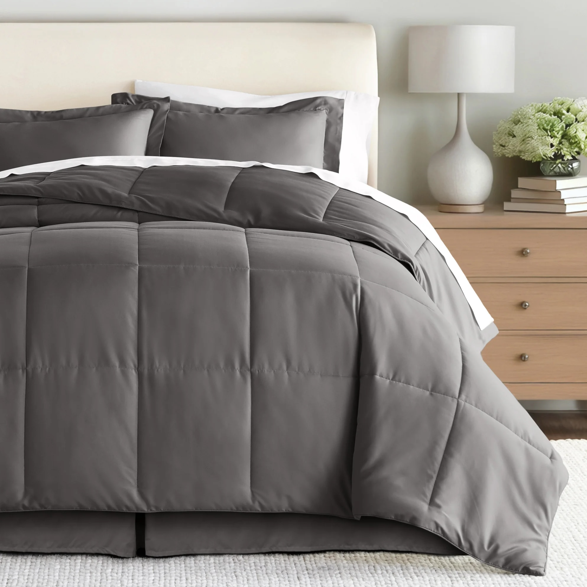 A neatly made bed features a gray, quilted comforter with matching pillow shams, set in a minimalist bedroom with a light wooden nightstand topped with books, a lamp, and a vase of flowers.