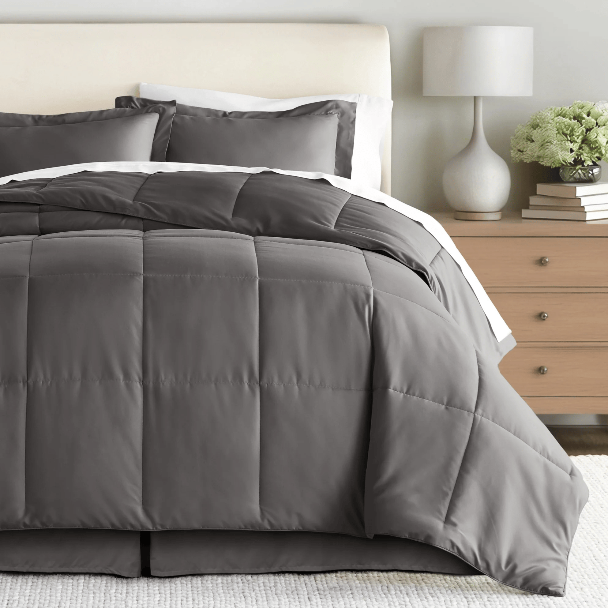 A neatly made bed features a gray, quilted comforter with matching pillow shams, set in a minimalist bedroom with a light wooden nightstand topped with books, a lamp, and a vase of flowers.