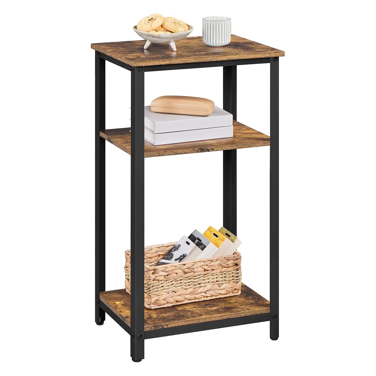 A three-tier storage end table with a rustic wood finish and black metal frame. The top shelf holds a bowl with cookies and a mug, the middle has books and a box, and the bottom has a wicker basket with items inside.