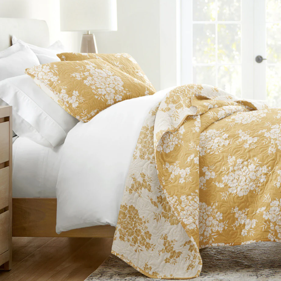 A bed with white linens features a gold and white floral patterned quilt and matching pillows, set in a bright room with a light wood nightstand and a beige lamp.