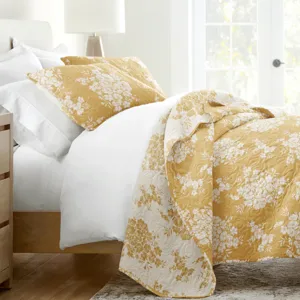 A bed with white linens features a gold and white floral patterned quilt and matching pillows, set in a bright room with a light wood nightstand and a beige lamp.