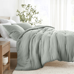 A bed is shown with a textured, sage green comforter and matching pillowcases, complemented by a wooden bedside table and a vase with greenery.