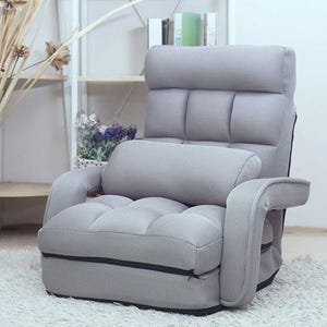 A modern, upholstered gray lounge chair with a high backrest, padded armrests, and a detachable cushion is placed on a white carpet, against a backdrop of a shelf with decorative items.