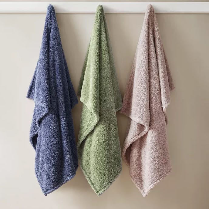 Three fluffy towels in blue, green, and pink are hanging on hooks against a light-colored wall.