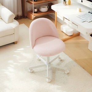 A pale pink upholstered swivel chair with white caster wheels sits in a bright, minimalist room next to a white desk, a white sofa, and a natural wood shelving unit.
