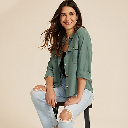 A woman wearing a relaxed-fit green shirt and distressed light blue jeans sits on a stool.