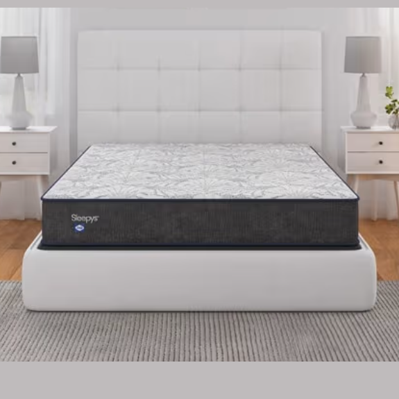 A mattress on a bed frame with a tufted headboard is flanked by two white nightstands with lamps, set on a wooden floor with a gray rug.