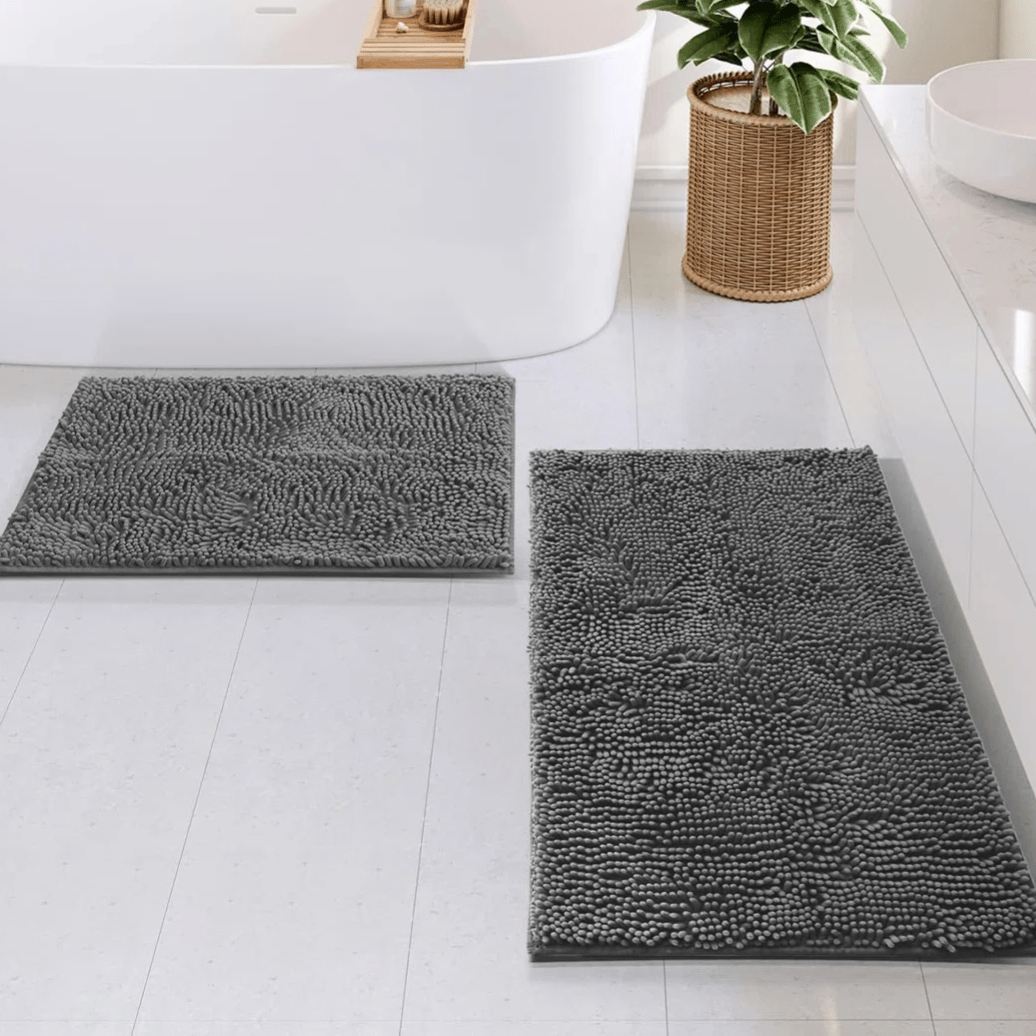 Two gray chenille bath mats of different sizes are placed on a light-colored bathroom floor, next to a freestanding bathtub and a woven plant basket.