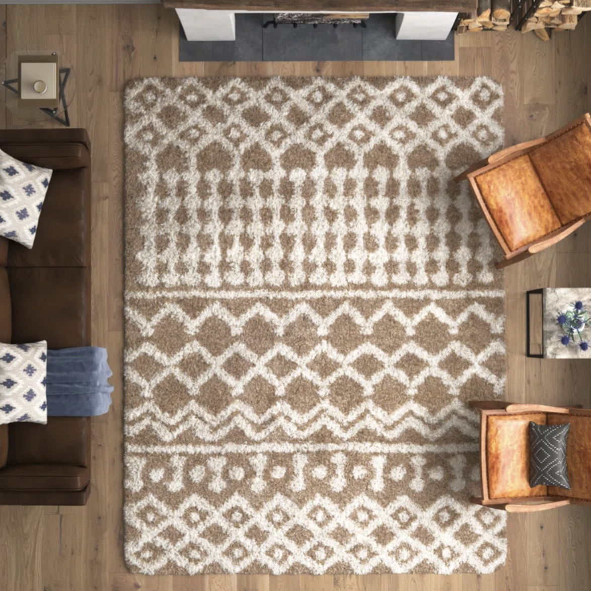 A room with a patterned beige and white area rug, a brown leather sofa with pillows, two brown armchairs, and a small side table with a potted plant. Wood flooring and a fireplace set the warm tone.