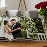 A wedding photo album open to a page showing a couple in wedding attire under a floral arch, placed on a wooden table next to a vase of red roses in a cozy living room setting.