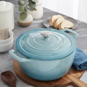 A light blue, enameled cast iron Dutch oven with a lid and handle, shown on a wooden surface, accompanied by a spoon, bowls, a vase with greenery, and a plate of sliced bread.