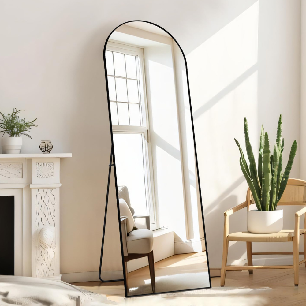 A freestanding, full-length arched mirror with a black frame is positioned in a bright room with a cactus in a white pot on a wooden chair and a white decorative fireplace mantel with a small plant and candle.