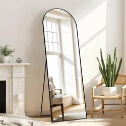 A freestanding, full-length arched mirror with a black frame is positioned in a bright room with a cactus in a white pot on a wooden chair and a white decorative fireplace mantel with a small plant and candle.
