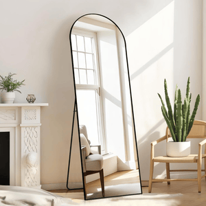 A freestanding, full-length arched mirror with a black frame is positioned in a bright room with a cactus in a white pot on a wooden chair and a white decorative fireplace mantel with a small plant and candle.
