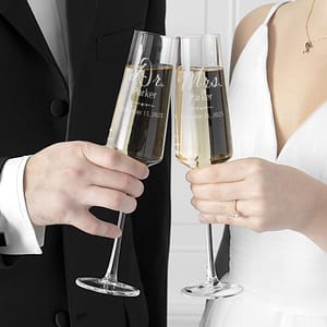 A set of two personalized champagne flutes, one engraved with \“Dr. Parker\“ and the other with \“Mrs. Parker,\“ dated September 15, 2025, held by a couple in formal attire toasting.