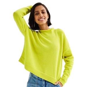 A woman is wearing a lime green long-sleeve sweater paired with blue jeans. She is smiling with one hand raised behind her head.
