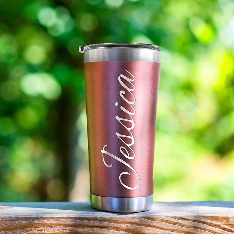 A metallic tumbler with a lid, featuring a pink gradient color and the name \“Jessica\“ written in an elegant white script font. It is designed for holding beverages and has an outdoor setting background.