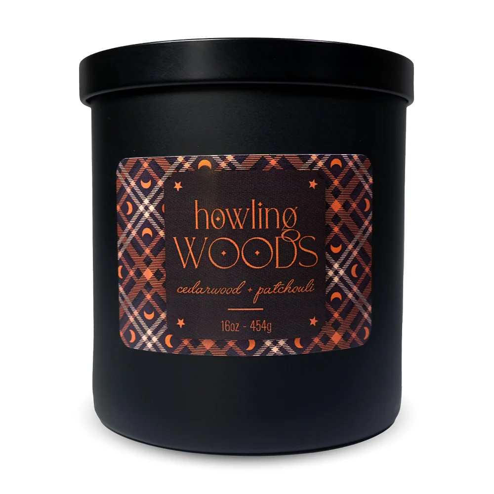 A black candle labeled \“howling WOODS\“ scented with cedarwood and patchouli, 16oz (454g), features an orange and black geometric pattern and moon design on the label.