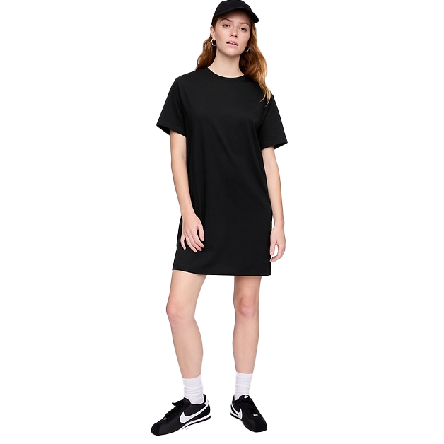 A woman is wearing a black T-shirt dress, paired with black sneakers and a black cap. She stands casually with one knee slightly bent.