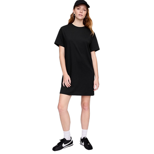A woman is wearing a black T-shirt dress, paired with black sneakers and a black cap. She stands casually with one knee slightly bent.