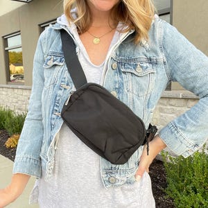A black nylon crossbody bag with a single adjustable strap is worn diagonally across the torso by a person in a denim jacket.