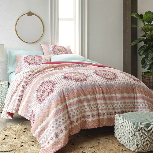 The Distant Lands Kora Bedding Set features a patterned comforter with a mix of geometric and floral designs in soft pinks, blues, and creams, paired with matching pillow shams. The set creates a cozy, bohemian look in a bedroom setting.