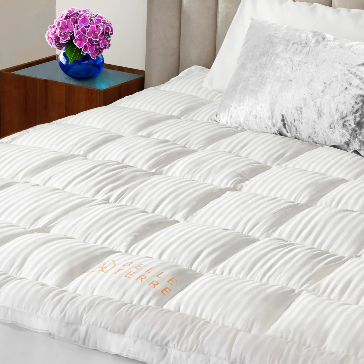 A white, quilted Queen Cooling Mattress Topper with a striped pattern, featuring the brand name \“Belle Terre.\“ It is topped with a gray pillow and accompanied by a bedside table with a blue vase holding pink flowers.