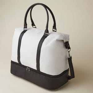 A two-tone weekender bag featuring a white upper portion and a dark base, with black straps and a detachable shoulder strap; the bag has a zippered closure on top.