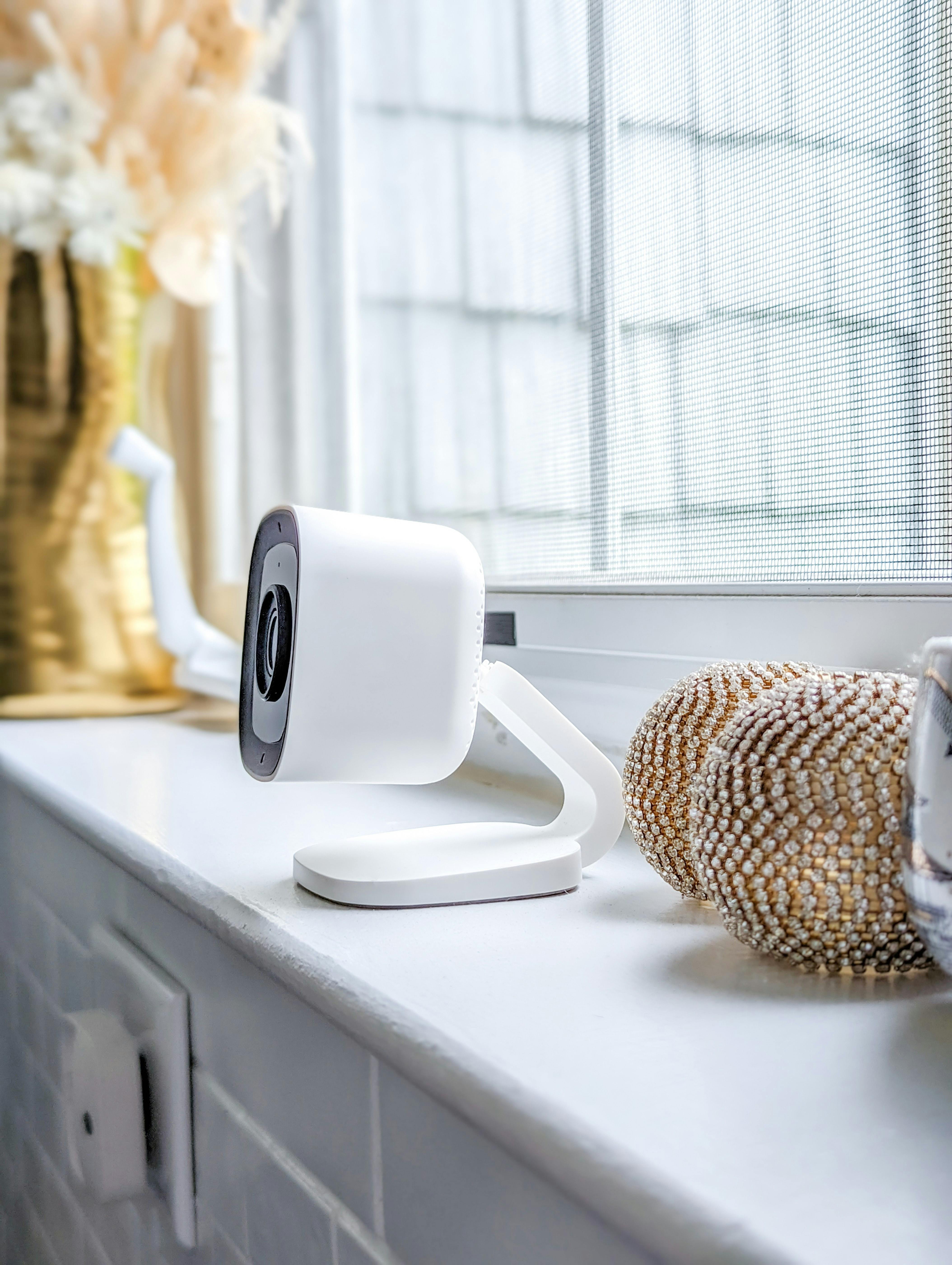 A white security camera with a flexible stand positioned on a windowsill next to a decorative sea urchin ornament.