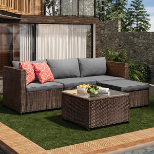 Outdoor patio set with a brown wicker sectional sofa featuring gray cushions and two red-patterned pillows, alongside a matching wicker coffee table topped with a glass surface, set on artificial grass and surrounded by greenery.