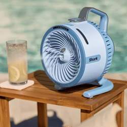 A blue Shark desk fan with a carry handle sits on a wooden table beside a glass of iced lemon water near a pool.