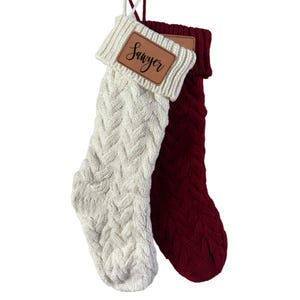 Two knit stockings hang together: one in cream with a personalized brown patch labeled \“Sawyer\“ and the other in deep red, both featuring a cable knit pattern.