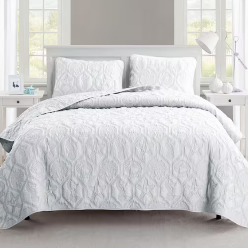 A neatly arranged bed features a white quilt with raised textures and matching pillow shams. The setting includes a bedside table with a lamp and decorative items, framed by windows.