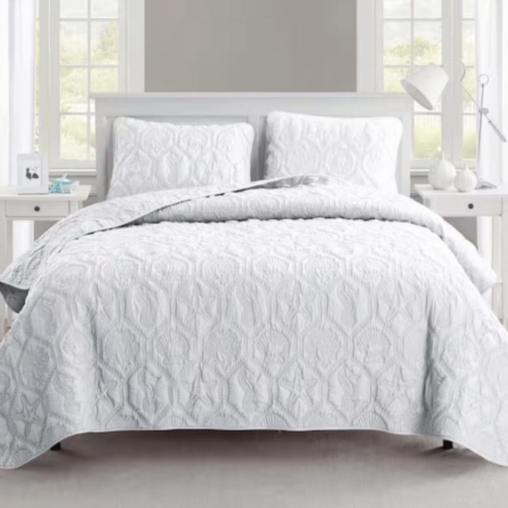 A neatly arranged bed features a white quilt with raised textures and matching pillow shams. The setting includes a bedside table with a lamp and decorative items, framed by windows.