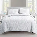 A neatly arranged bed features a white quilt with raised textures and matching pillow shams. The setting includes a bedside table with a lamp and decorative items, framed by windows.