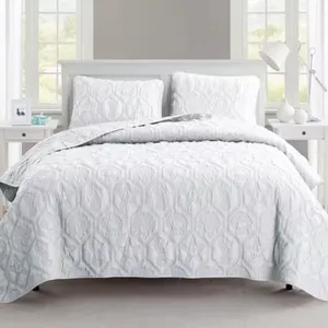 A neatly arranged bed features a white quilt with raised textures and matching pillow shams. The setting includes a bedside table with a lamp and decorative items, framed by windows.