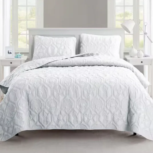 A neatly arranged bed features a white quilt with raised textures and matching pillow shams. The setting includes a bedside table with a lamp and decorative items, framed by windows.