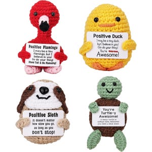 Four crocheted plush toys: a red flamingo, yellow duck, brown and white sloth, and green turtle. Each holds a sign with positive affirmations.