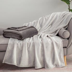 Two plush Velvetsoft throws are draped over a couch, one in gray neatly folded and the other in white casually spread out.