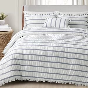 A bed is styled with a white comforter featuring thin gray stripes and ruffled edges, accompanied by matching pillows. A small plant and books sit on a wicker side table.