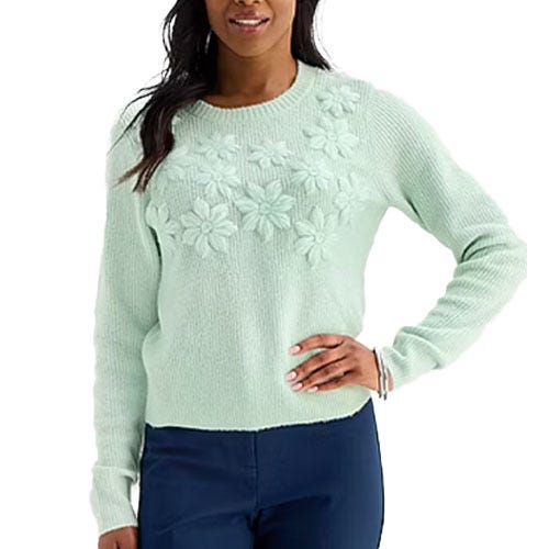 A woman wears a light mint green sweater with floral embroidery across the chest, paired with dark blue pants.
