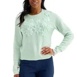 A woman wears a light mint green sweater with floral embroidery across the chest, paired with dark blue pants.