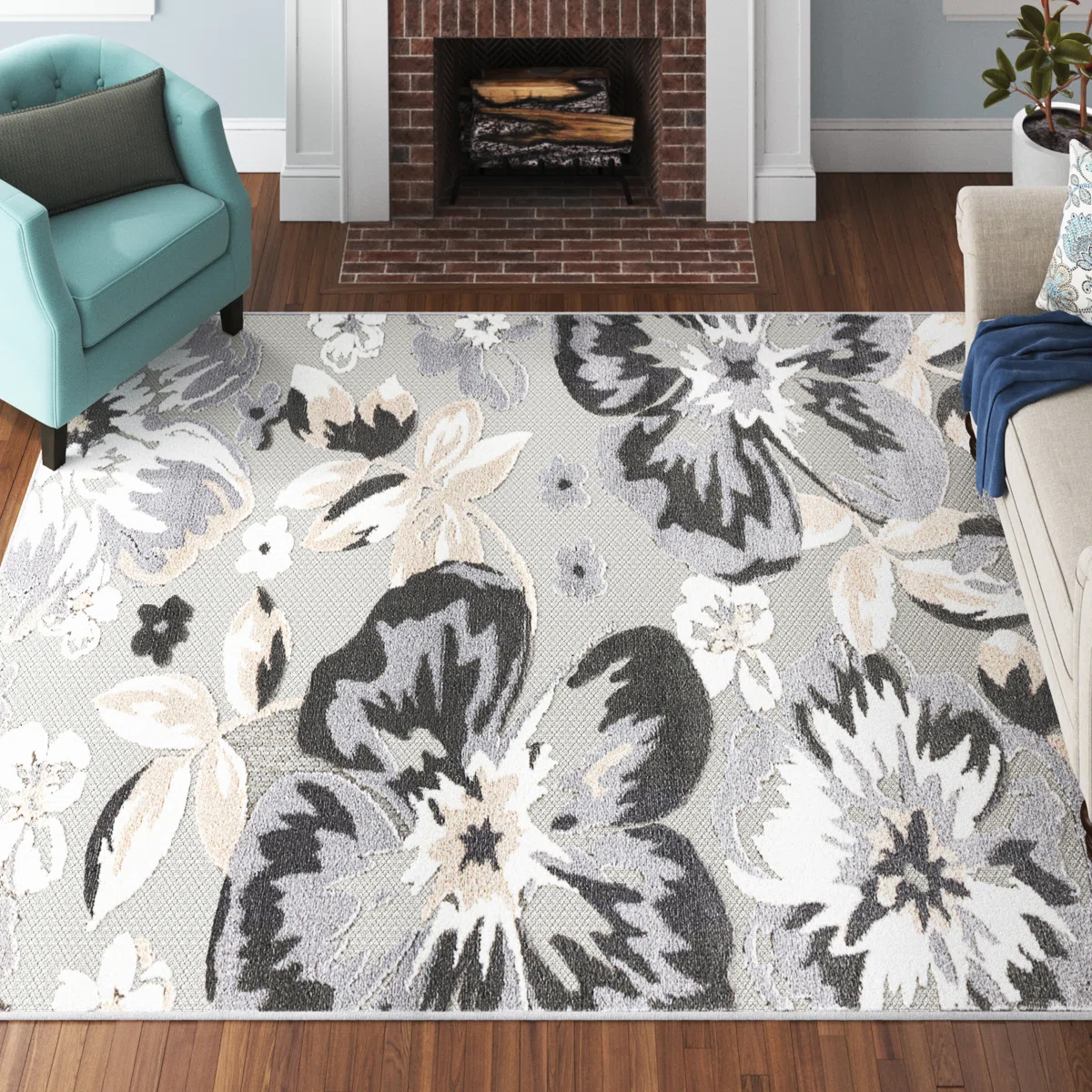 A living room features a large area rug with a floral pattern in shades of gray, white, and beige, set on wooden flooring. An aqua armchair and light-colored sofa flank a brick fireplace.